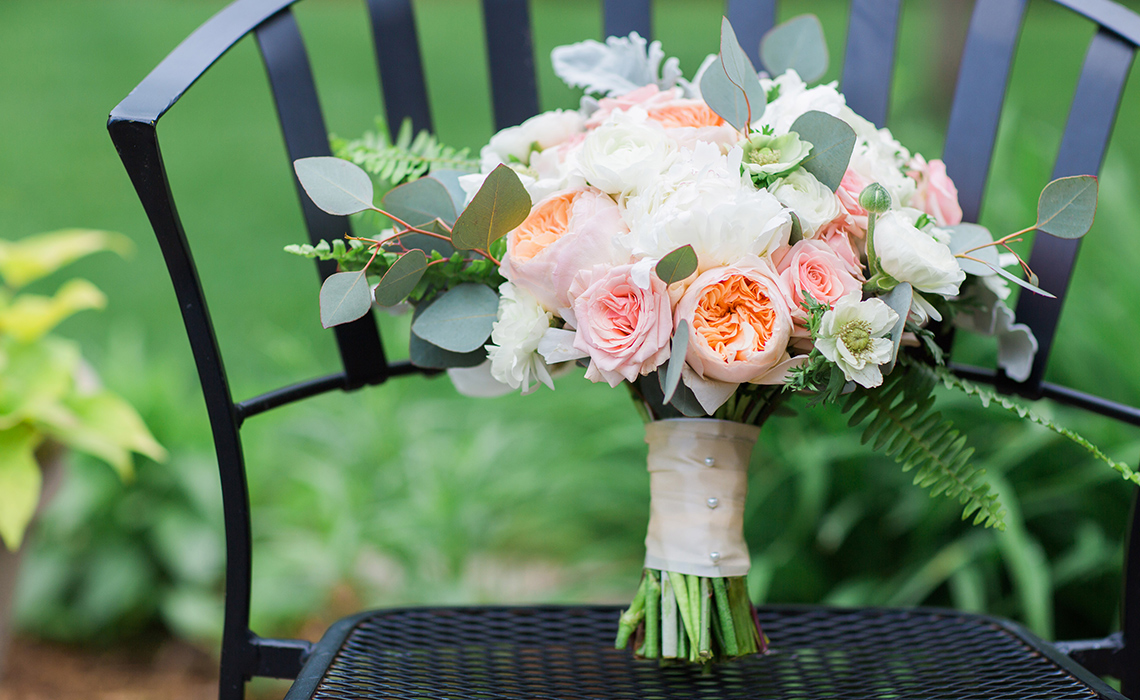 Maureen's bridal bouquet, by A Moment in Time, rest on a chair outdoors before their wedding ceremony in Farmington Hills, Michigan.